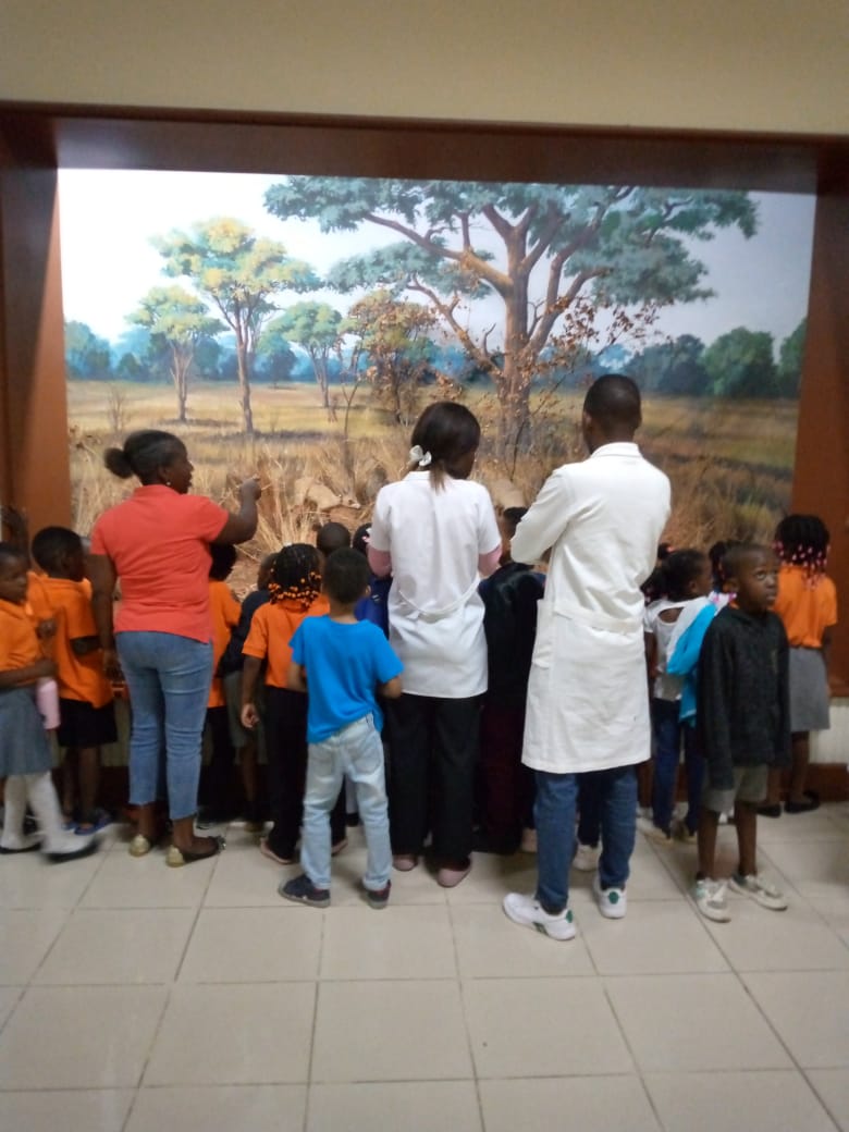 Crianças e professores numa visita a um museu, diante de um painel de paisagem